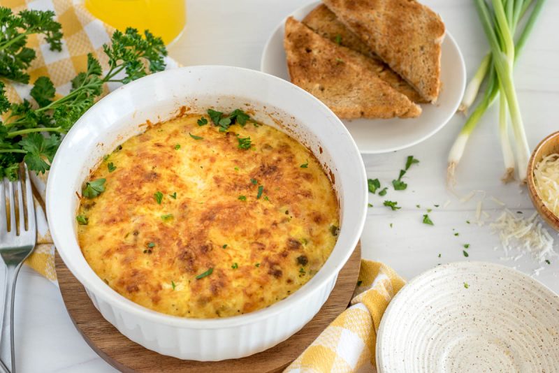 the finished and browned ham and cheese quiche in a white soufflé plan with toast and parsley in the background