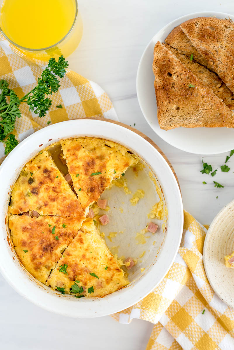 An overhead vertical shot of a white soufflé pan filled with slices of ham and egg quiche, with toast and a yellow and white checkered napkin