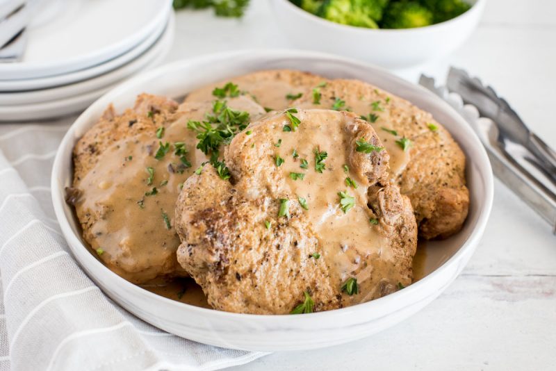 close up on a white bowl with four pork chops in mushroom gravy with steamed broccoli in the background