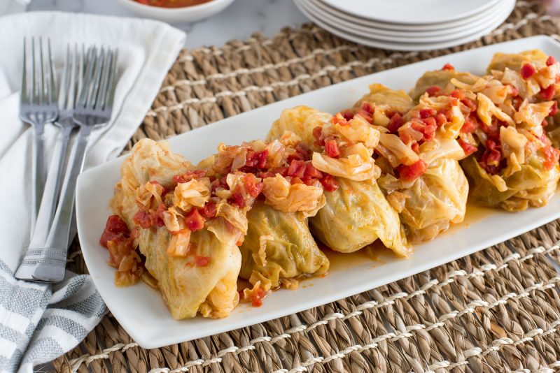 Stuffed Cabbage Rolls in a row, with red tomatoes and chopped cabbage in a sauce on top of the rolls, served on a white plate, with white napkins and a brown woven placemat underneath
