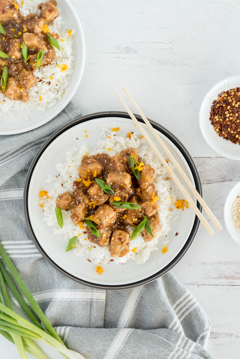 an overhead shot of a plate of orange chicken, served over a bed of rice and garnished with green onions. A pair of chopsticks are at the top right side of the plate