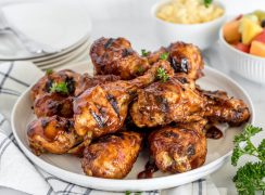 A white plate on a napkin with a pile of BBQ chicken legs on top, with a white cup of fruit salad in the background