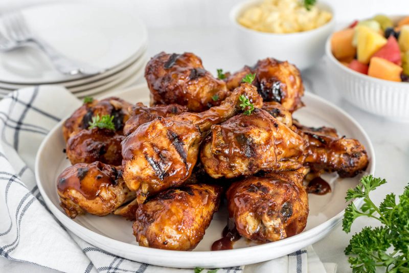 A white plate on a napkin with a pile of BBQ chicken legs on top, with a white cup of fruit salad in the background