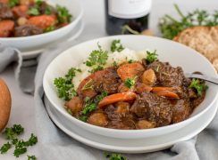 Close up picture of Instant Pot beef bourguignon served with mashed potatoes and fresh parsley in a white bowl with rolls and a bottle of red wine in the background.