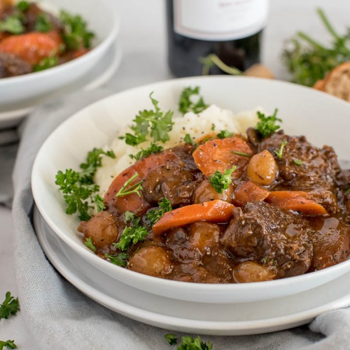 Close up picture of Instant Pot beef bourguignon served with mashed potatoes and fresh parsley in a white bowl with rolls and a bottle of red wine in the background.