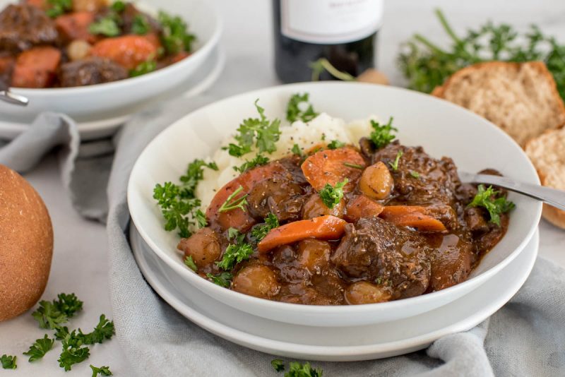 Close up picture of Instant Pot beef bourguignon served with mashed potatoes and fresh parsley in a white bowl with rolls and a bottle of red wine in the background.