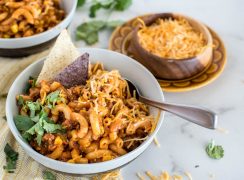 Close-up shot of Instant Pot Chili Pasta, served in a white bowl and garnished with cilantro, shredded cheese, and white and blue corn chips, with a bowl of shredded cheese in the background