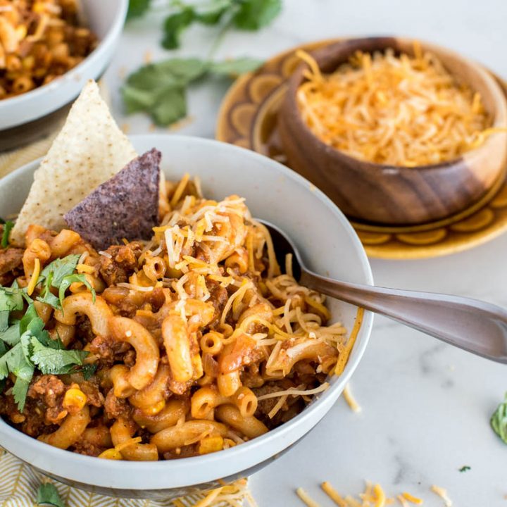 Close-up shot of Instant Pot Chili Pasta, served in a white bowl and garnished with cilantro, shredded cheese, and white and blue corn chips, with a bowl of shredded cheese in the background