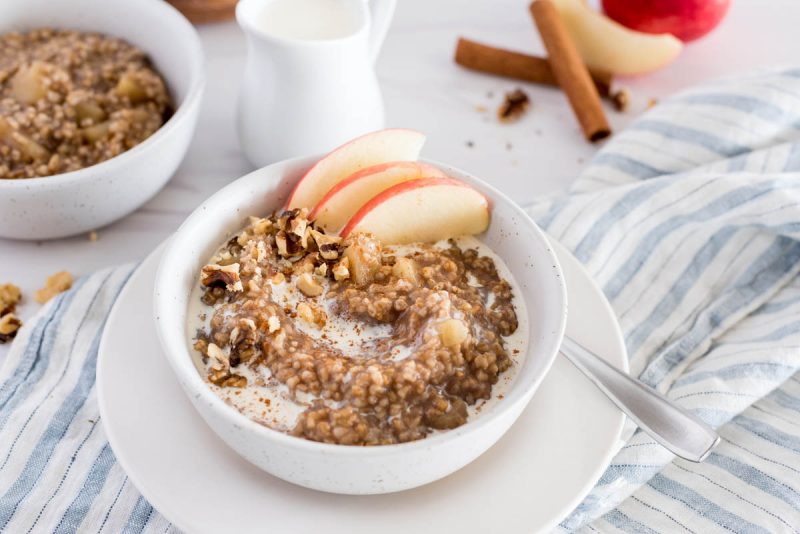 white bowl of apple-cinnamon flavored instant pot steel cut oats with apple slices, on top of a muted blue and white striped napkin with a glass of milk and a second bowl of steel cut oats in the background