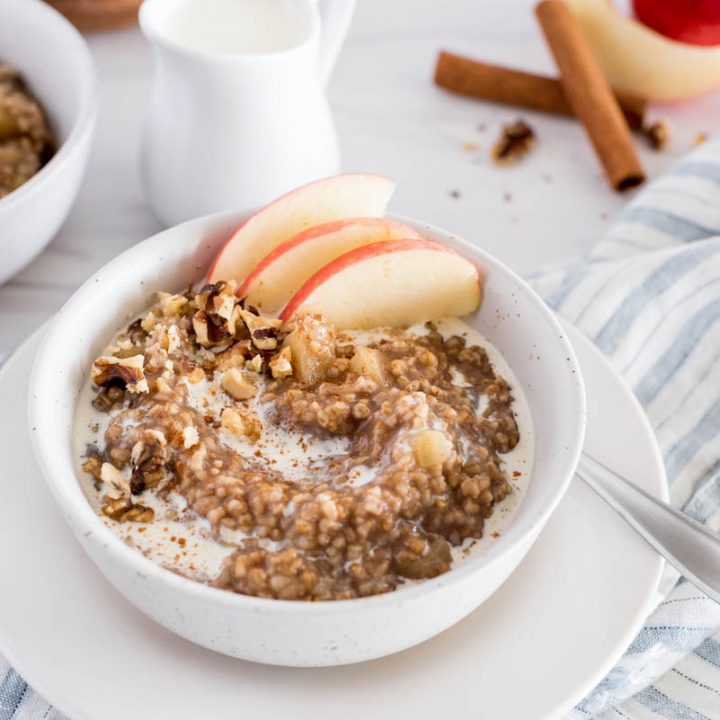 white bowl of apple-cinnamon flavored instant pot steel cut oats with apple slices, on top of a muted blue and white striped napkin with a glass of milk and a second bowl of steel cut oats in the background