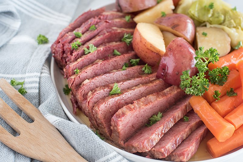 45 degree shot of thin slices of Instant Pot corned beef served with tender carrots, white potatoes, and cabbage, garnished with fresh parsley on a round white serving dish