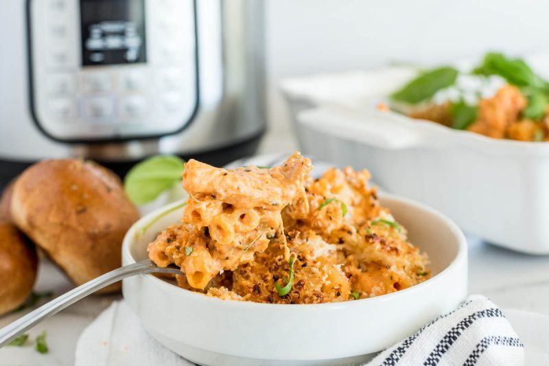 A horizontal shot of a small white bowl filled with cooked ziti and a spoon, sitting in front of an Instant Pot and several rolls