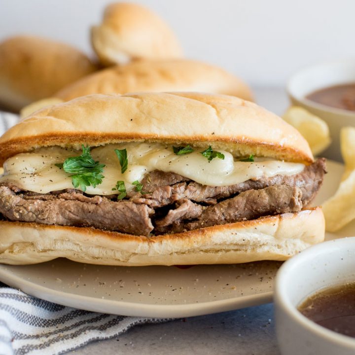 A close up of an Instant Pot French dip sandwich topped with cheese on a bun next to a bowl of au jus.