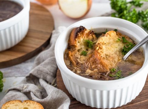 A finished serving of Instant Pot French Onion soup, with browned bread and melted cheese on top of a rich brown french onion soup., served in a white ramekin with a second bowl in the background