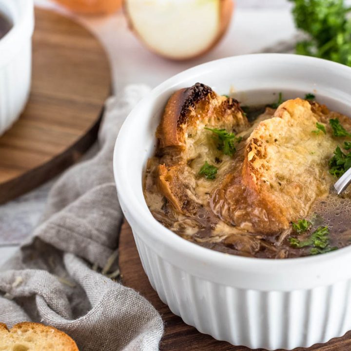 A finished serving of Instant Pot French Onion soup, with browned bread and melted cheese on top of a rich brown french onion soup., served in a white ramekin with a second bowl in the background