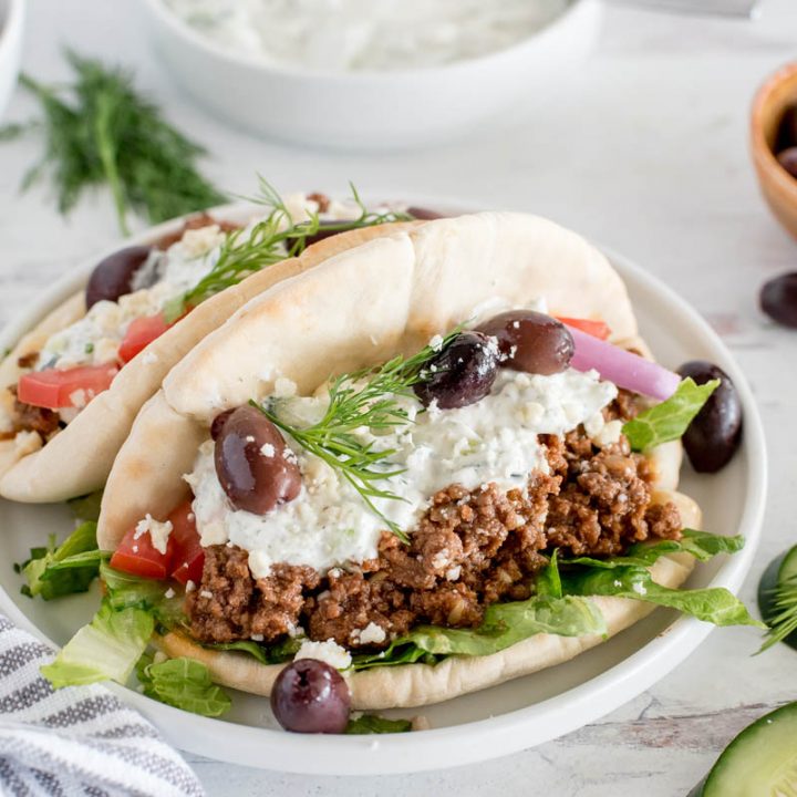 plate with two greek beef tacos topped with tzatziki sauce, olives, cucumbers and dill