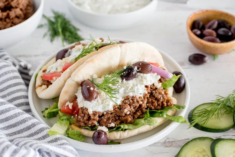 plate with two greek beef tacos topped with tzatziki sauce, olives, cucumbers and dill