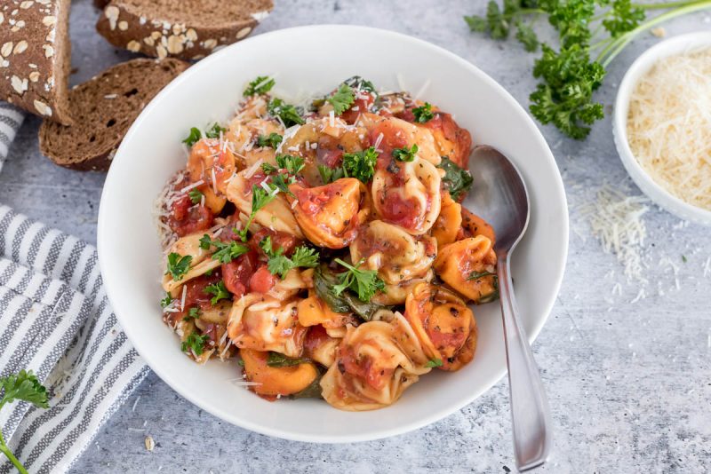 Close up picture of Instant Pot tortellini topped with fresh parmesan and parsley, placed in a white bowl. Additional parmesan and slices of wheat bread in the background.
