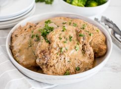 close up on a white bowl with four pork chops in mushroom gravy with steamed broccoli in the background