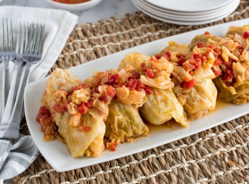 Stuffed Cabbage Rolls in a row, with red tomatoes and chopped cabbage in a sauce on top of the rolls, served on a white plate, with white napkins and a brown woven placemat underneath