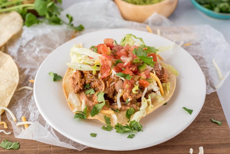 45 degree close up of a tostada, all plated up on a white plate on a yellow corn tortilla, topped with sweet pork, tomatoes, and cilantro.