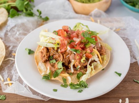 45 degree close up of a tostada, all plated up on a white plate on a yellow corn tortilla, topped with sweet pork, tomatoes, and cilantro.