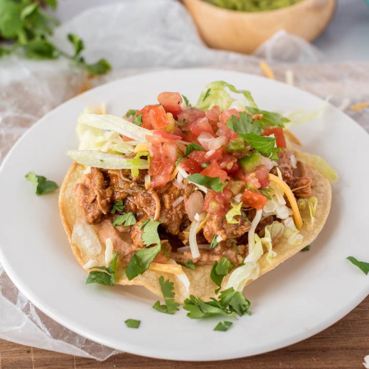 45 degree close up of a tostada, all plated up on a white plate on a yellow corn tortilla, topped with sweet pork, tomatoes, and cilantro.