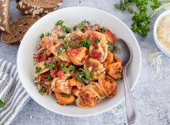 Close up picture of Instant Pot tortellini topped with fresh parmesan and parsley, placed in a white bowl. Additional parmesan and slices of wheat bread in the background.