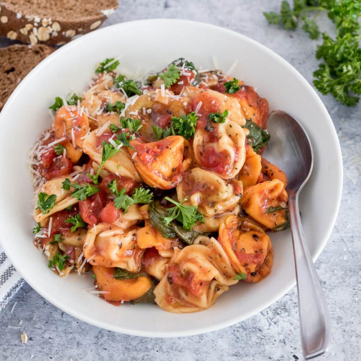 Close up picture of Instant Pot tortellini topped with fresh parmesan and parsley, placed in a white bowl. Additional parmesan and slices of wheat bread in the background.