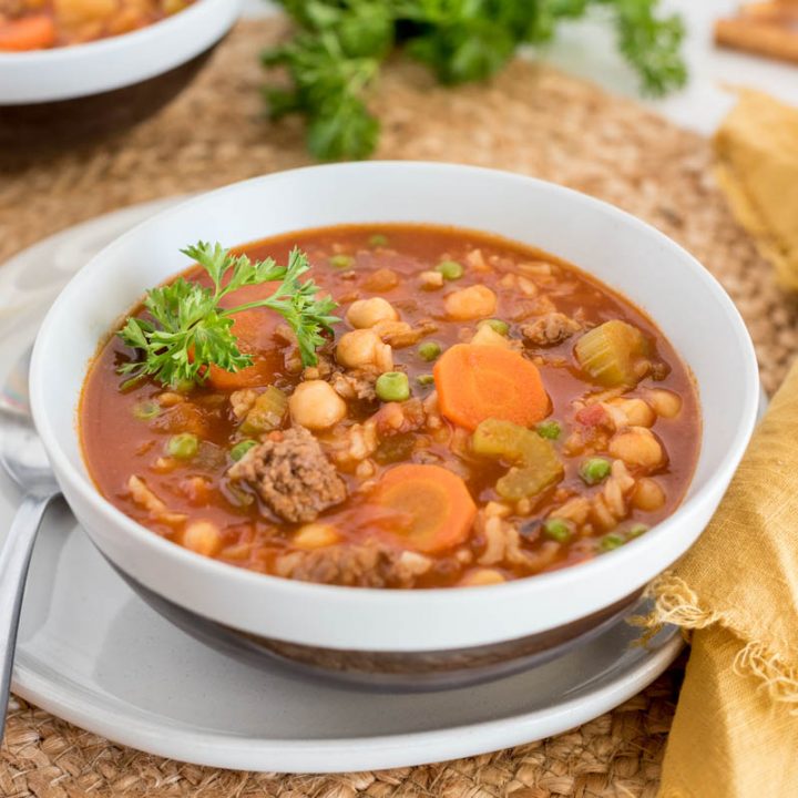 Close up picture of Instant Pot vegetable beef and rice soup served in a white bowl. Placed on a woven mat, next to a yellow linen napkin.