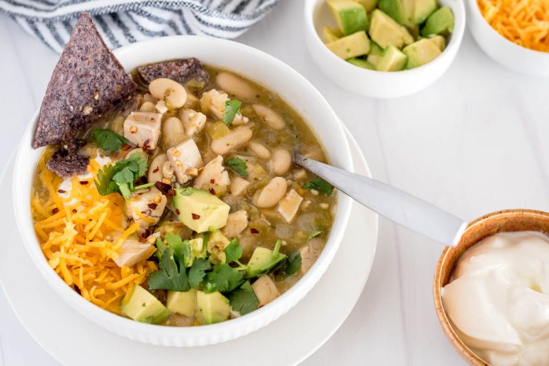 bowl of instant pot white chicken chili with avocado, chips and shredded cheese on top