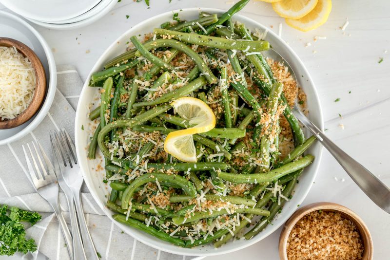 overhead shot of a white bowl of green beans with a lemon twist garnish and breadcrumbs