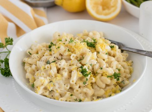 a 45 degree close-up shot of a white bowl filled with creamy lemon pasta, with additional parsley and lemon for garnish, and a old silver fork at the top right