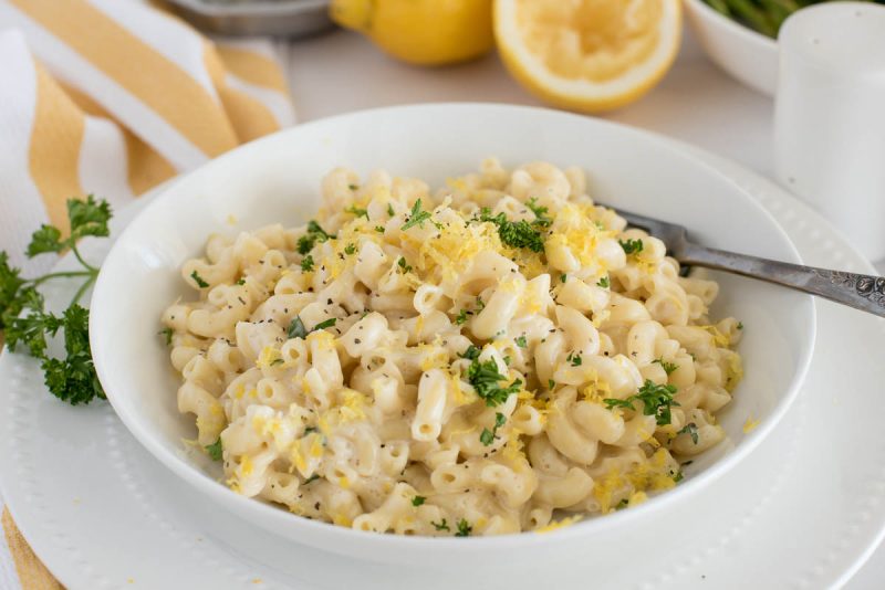 a 45 degree close-up shot of a white bowl filled with creamy lemon pasta, with additional parsley and lemon for garnish, and a old silver fork at the top right