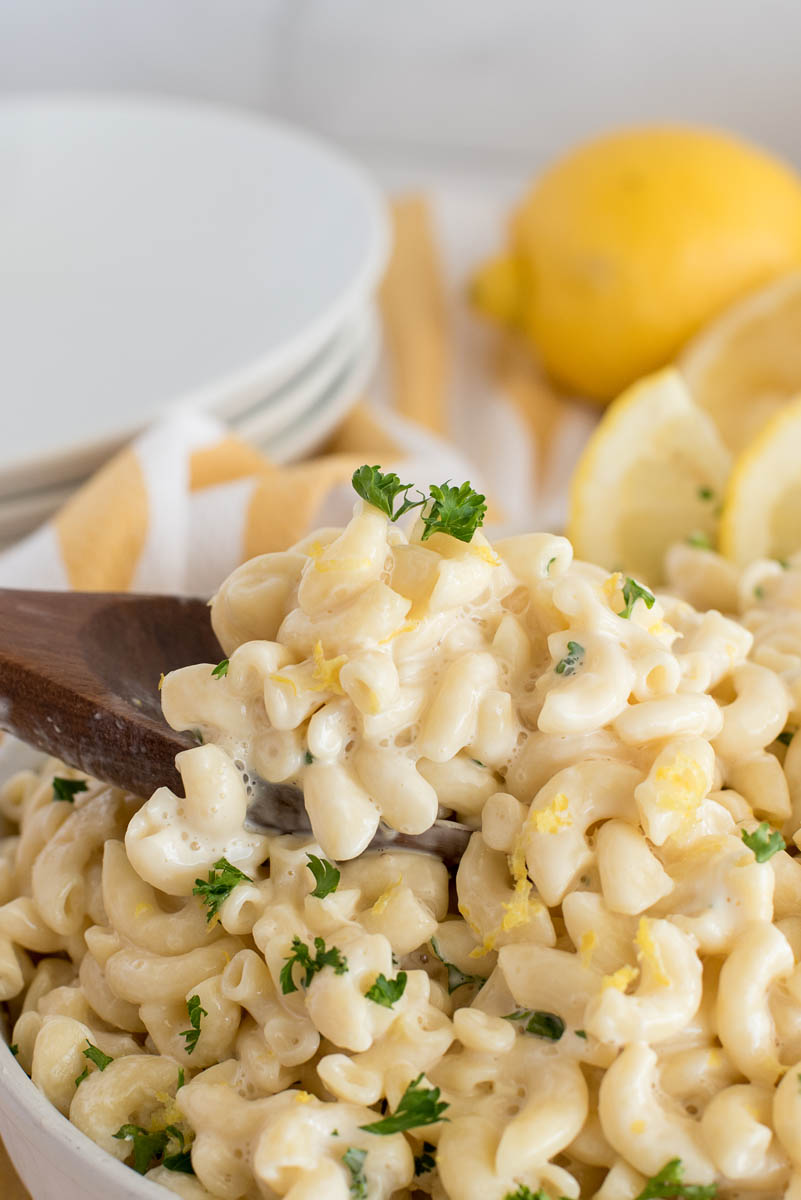 a close-up of a dark brown wooden spoon scooping up a serving of creamy lemon pasta, with a stack of white plates and golden-yellow lemons in the background