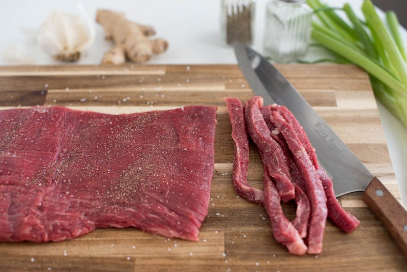 thinly sliced flank steak on a wooden cutting board with ginger and garlic and onions in the background