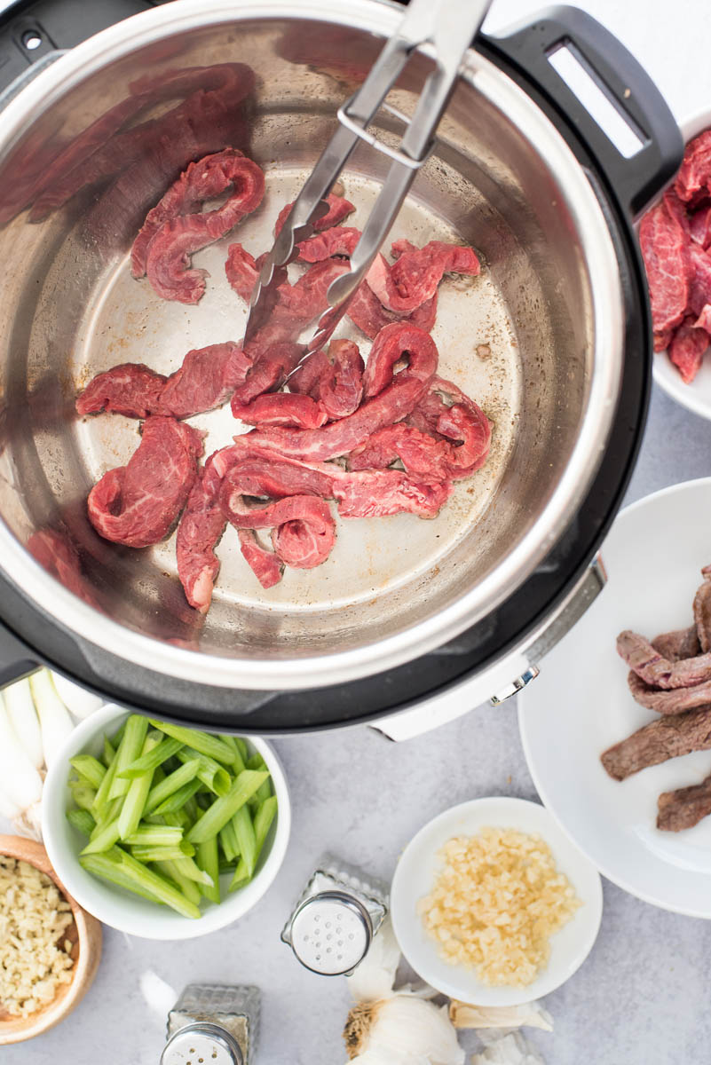 an overhead shot of flank steak sauteeing in an Instant Pot with the other food ingredients in bowls to the lower portion of the photo