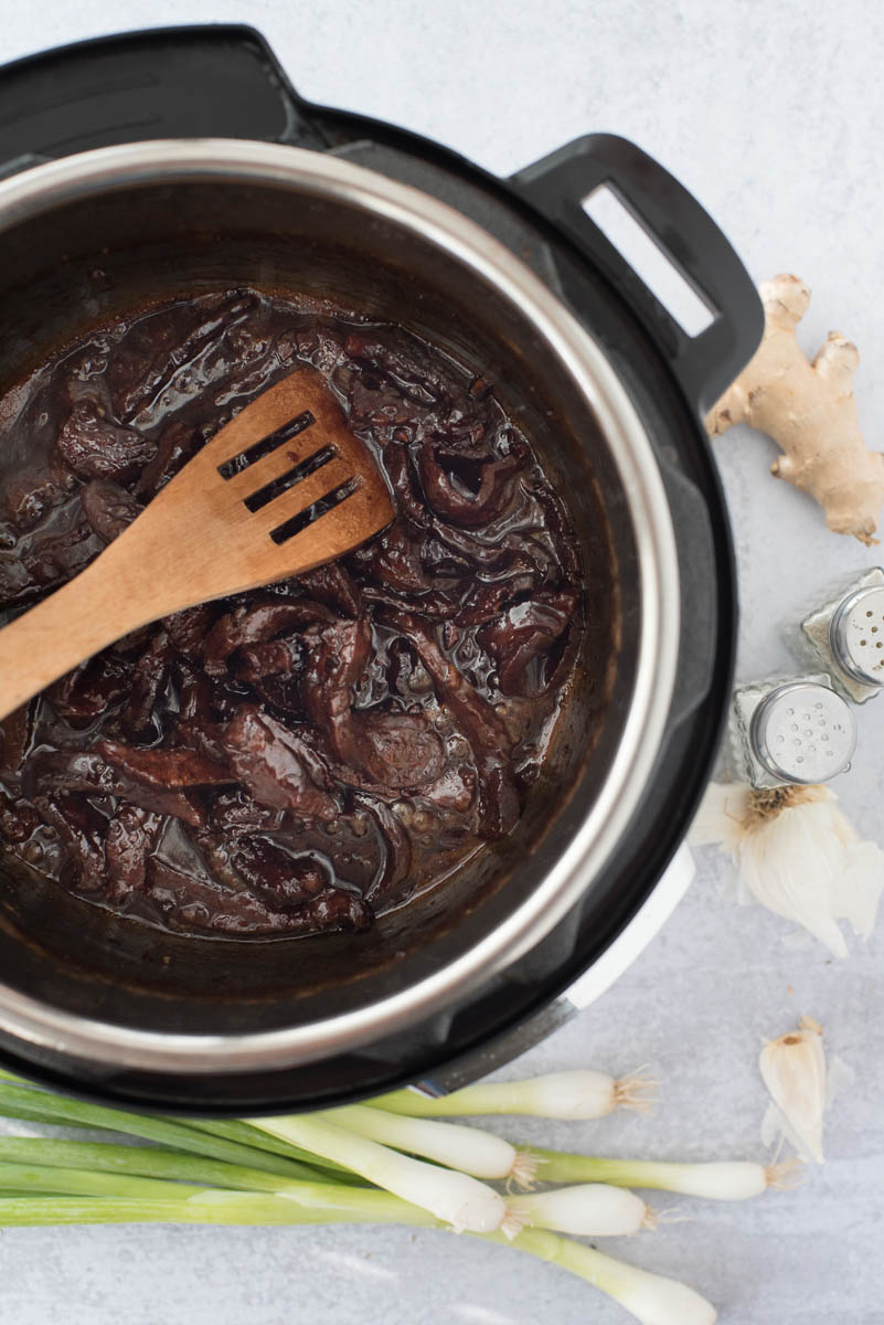 an overhead shot of mongolian beef cooked and in a thick sauce inside the Instant Pot with a wooden spoon stirring them and green onions on the bottom of the photo