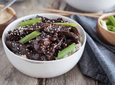 a white bowl that's close-up featuring thin cut strips of beef flank steak served over white rice with sesame seeds and green onions