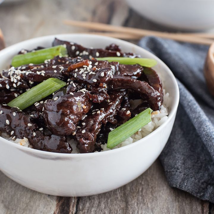 a white bowl that's close-up featuring thin cut strips of beef flank steak served over white rice with sesame seeds and green onions