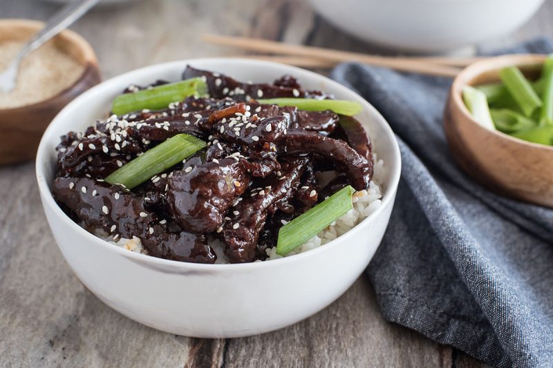 a close-up of a white bowl filled with mongolian beef—thinly cut flank steak cooked in the Instant Pot and 
coated in a dark sauce with green onions and sesame seeds for garnish