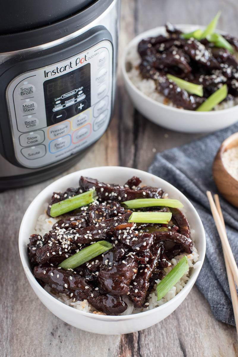 an overhead shot showing a white bowl of cooked Instant Pot Mongolian Beef garnished with scallions and sesame seeds in front of an Instant Pot with a second bowl of pressure cooked mongolian beef in the background