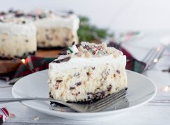 a close-up shot of a slice of Instant Pot Cheesecake on a white plate with a fork in the foreground. The cheesecake has clearly visible peppermint bark chips throughout the creamy white cheesecake layer