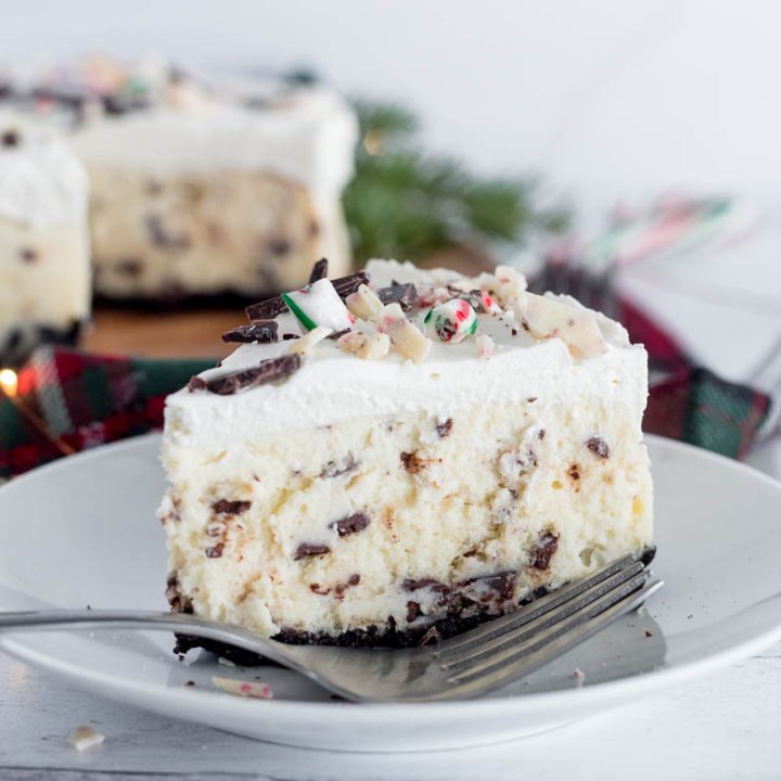 a close-up shot of a slice of Instant Pot Cheesecake on a white plate with a fork in the foreground. The cheesecake has clearly visible peppermint bark chips throughout the creamy white cheesecake layer