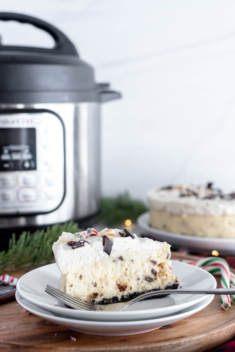 a head-on shot of a slice of peppermint cheesecake on a white plate in front of in the Instant Pot  and evergreen boughs and candy canes