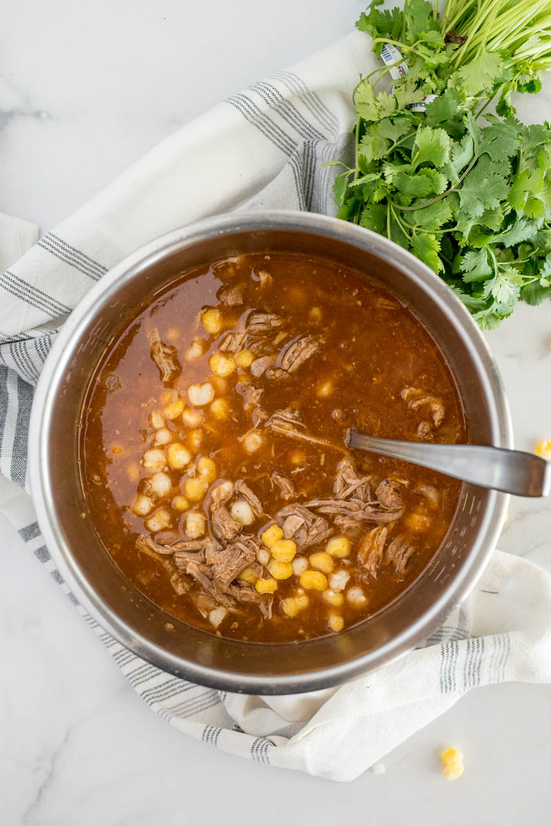 The cooking pot of a pressure cooker filled with the finished pork and hominy stew (posole), with a napkin, cilantro, and hominy in the background