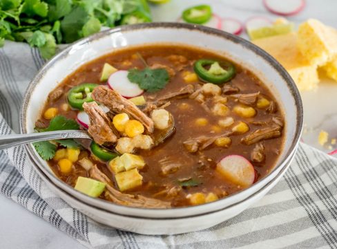 a horizontal 45 degree shot of a spoonful of InstaPot Pork Stew with shredded pork, hominy, avocado,, and a slice of cornbread and radishes in the background