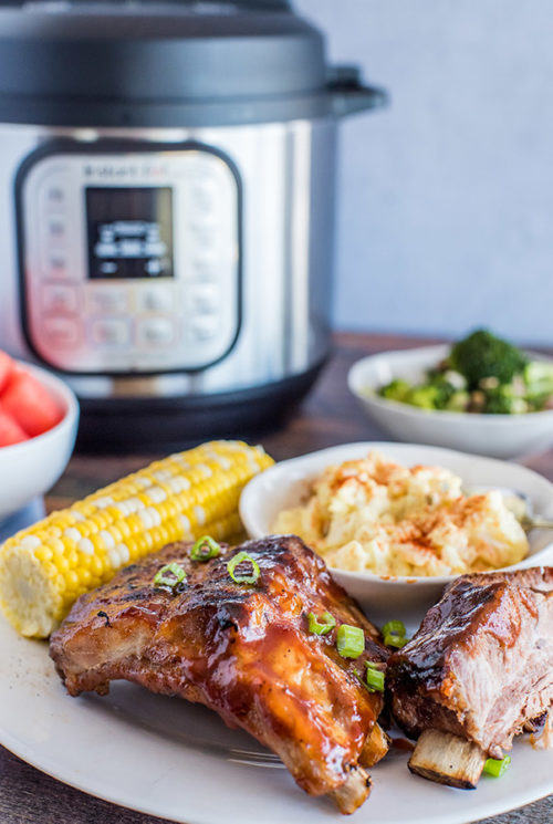 A plate of food on a table with Pork ribs, corn on the cob, and potato salad with Instant Pot in background.