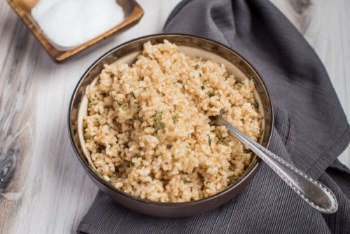 A dark brown bowl full of short-grain brown rice, prepared in the pressure cooker, with a garnish of sea salt in the background