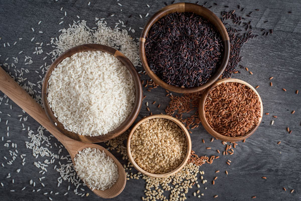 An overhead shot of four wooden bowls filled with different types of rice—white, brown, black, and pink—with additional white rice in a wooden spoon